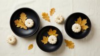 Autumn flat lay with white pumpkins and golden leaves in black bowls on neutral background for seasonal home decor and thanksgiving styling.