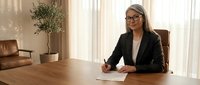 Professional businesswoman in glasses sitting at wooden desk in modern office with natural lighting and leather furniture for corporate presentations.