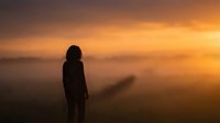 Silhouette of woman standing in misty landscape during golden hour sunrise, contemplative mood with warm orange sky and fog creating atmospheric scene.