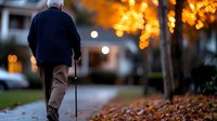 Senior man with walking cane strolling along autumn path with fallen leaves at dusk, illuminated by warm bokeh lights, creating peaceful retirement scene.