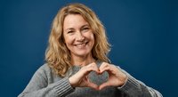 Happy middle-aged Caucasian woman making heart shape with hands wearing gray sweater on blue background expressing love and positivity for lifestyle content.