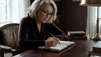 Senior Caucasian woman with gray hair and glasses writing in notebook at wooden desk in home office, focused on paperwork and planning tasks.