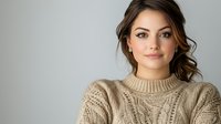 Young Caucasian woman with brown hair and natural makeup wearing beige knit sweater with cable pattern, looking at camera with gentle smile against light gray background.