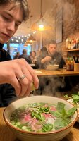 Young person with light hair and fair complexion preparing Asian noodle soup with fresh herbs and meat in casual restaurant setting with warm lighting and blurred diners in background.