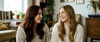 Two young women friends laughing together on couch in cozy living room wearing cream sweaters during casual conversation at home.