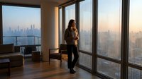 Young woman with dark hair and medium skin tone, wearing business casual attire, standing by floor-to-ceiling windows in high-rise office overlooking city skyline at sunset, contemplating urban view.