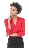 Young woman with natural curly hair in red blouse and black skirt looking thoughtful with hand on chin, isolated on white background.