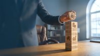 Businessman placing a security shield on a tower of wooden blocks with finance and real estate icons, symbolizing asset protection and investment safety.