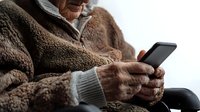 Senior Caucasian person in cozy brown fleece jacket using smartphone, close-up view of aged hands holding mobile device while sitting in comfort.