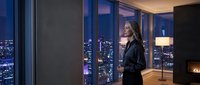 Professional businesswoman in elegant black suit standing by floor-to-ceiling windows overlooking illuminated city skyline at night in modern luxury apartment.