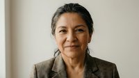 Professional mature Hispanic businesswoman in elegant suit smiling confidently in modern office environment for corporate leadership and executive success concepts.