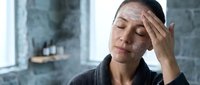 Young Asian woman experiencing headache or stress touching forehead with eyes closed in modern bathroom setting for wellness and healthcare concepts.
