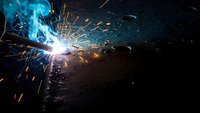 Industrial welding process with bright electric arc and flying sparks on metal surface in manufacturing workshop environment.