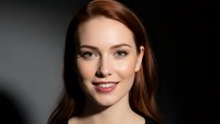 Young Caucasian woman with auburn hair smiling warmly at camera against dark background. Professional headshot for business and lifestyle content.