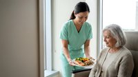 Asian female healthcare worker in green scrubs serving nutritious meal to elderly Caucasian woman patient in bright medical facility room for senior care services.