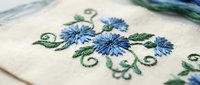 Blue cornflower embroidery on cream fabric with green leaves and decorative swirls. Traditional needlework craft for home decor and textile projects.