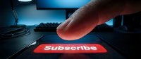 Finger pressing glowing red subscribe button on smartphone screen in dark room with computer setup in background.