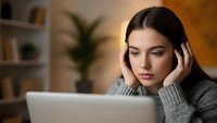 Young Caucasian woman experiencing stress while working on laptop computer in modern home office environment with warm lighting and plants.