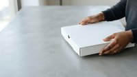 Person with dark skin tone wearing black long-sleeve shirt holding white box on gray concrete surface, suitable for e-commerce, packaging, delivery, and product presentation concepts.