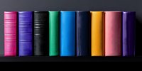 Colorful hardcover books arranged in row on black shelf, spines showing vibrant pink, purple, black, mint, blue, orange and violet colors creating minimalist composition.