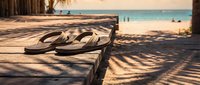 Flip-flops on wooden deck overlooking tropical beach with turquoise water and palm tree shadows at sunset for vacation and summer relaxation concept.