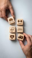 Hands arranging wooden blocks with healthcare icons including meditation lotus, fitness dumbbell, stethoscope, medical cross and ambulance symbols.