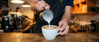 Professional barista creating latte art by pouring steamed milk into coffee cup at modern cafe counter with warm ambient lighting and coffee equipment in background.