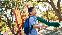 Happy Asian elementary school boy with backpack playing on colorful playground equipment during sunny day at park for outdoor recreation and childhood fun.