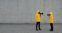 Two people in bright yellow hoodies holding megaphones facing each other against concrete wall background for communication and protest concepts.