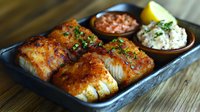 Grilled cod fillets with crispy seasoned crust served on black plate with two dipping sauces and lemon wedge garnish, fresh herbs sprinkled on top for restaurant menu.