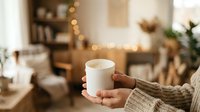 Hands holding warm white mug in cozy home interior with holiday decorations, bokeh lights and wooden furniture for seasonal comfort and relaxation themes.