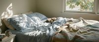 Unmade bed with rumpled blue and white bedding, natural sunlight streaming through window, cozy bedroom interior with casual, lived-in aesthetic.