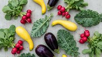 Fresh organic vegetables including kale, radishes, yellow squash and eggplant arranged on neutral background for healthy cooking and nutrition concepts.