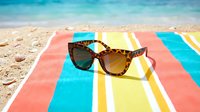 Stylish tortoiseshell sunglasses on colorful striped beach towel with ocean waves and sandy shore in background for summer vacation and travel concepts.