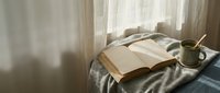 Cozy morning scene with open book and coffee cup on unmade bed near window with soft natural light creating peaceful reading atmosphere for relaxation.