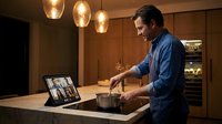 Man in blue shirt having video conference call on tablet at wooden desk with pendant lights, suitable for remote work, business communication, and virtual meeting concepts.