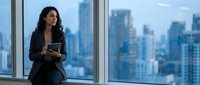 Professional Hispanic businesswoman with tablet standing by office window overlooking city skyline during daytime for corporate presentations.