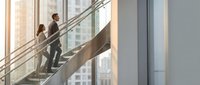 Two professional businesspeople climbing modern office staircase in corporate building with glass windows and city view during daytime for career advancement concept.