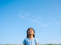 Young girl with dark hair and medium skin tone wearing light blue shirt, looking upward with joyful expression against clear blue sky and grass field, conveying happiness and freedom.