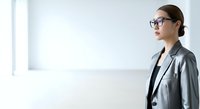 Young East Asian woman wearing glasses and gray blazer standing in bright, minimalist office space, suitable for corporate, business, and professional lifestyle content.