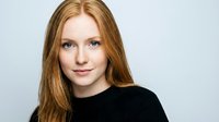 Young Caucasian woman with long red hair and blue eyes wearing black top smiling at camera against neutral background for professional headshot.