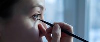 Young woman applying black eyeliner makeup with precision brush near bright window. Professional beauty routine and cosmetic application concept.