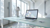 Modern minimalist office workspace with laptop computer on glass desk near large windows with natural light and contemporary decor elements.