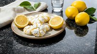 Fresh lemon crinkle cookies with powdered sugar on wooden plate alongside whole lemons and glass of milk on dark granite countertop for baking content.