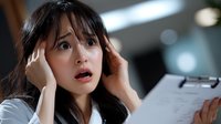 Young Asian businesswoman looking stressed and overwhelmed while holding document, expressing shock and anxiety at workplace with hands on head.
