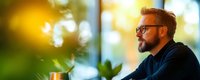 Thoughtful bearded businessman with glasses looking out office window during golden hour, contemplating future plans and strategies in modern workplace.