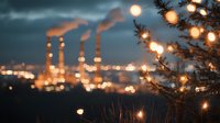 Industrial factory chimneys at dusk with glowing lights and smoke stacks creating atmospheric bokeh effect through tree branches in foreground, moody evening scene.