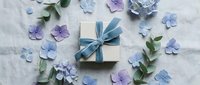 Gift box with blue ribbon surrounded by purple and blue paper flowers on white linen background for celebrations and special occasions.