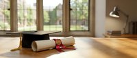 Graduation cap and diploma with red ribbon on wooden floor in bright room with large windows, symbolizing academic achievement and educational success.