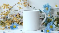White ceramic mug mockup surrounded by delicate blue daisies and yellow dried flowers on white background, styled product photography for branding and design.